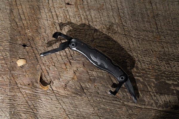 A black FLE Pocket Jack 2.0 woodcarving multi-tool with several partially extended blades rests on a rough, weathered wooden surface in natural light.