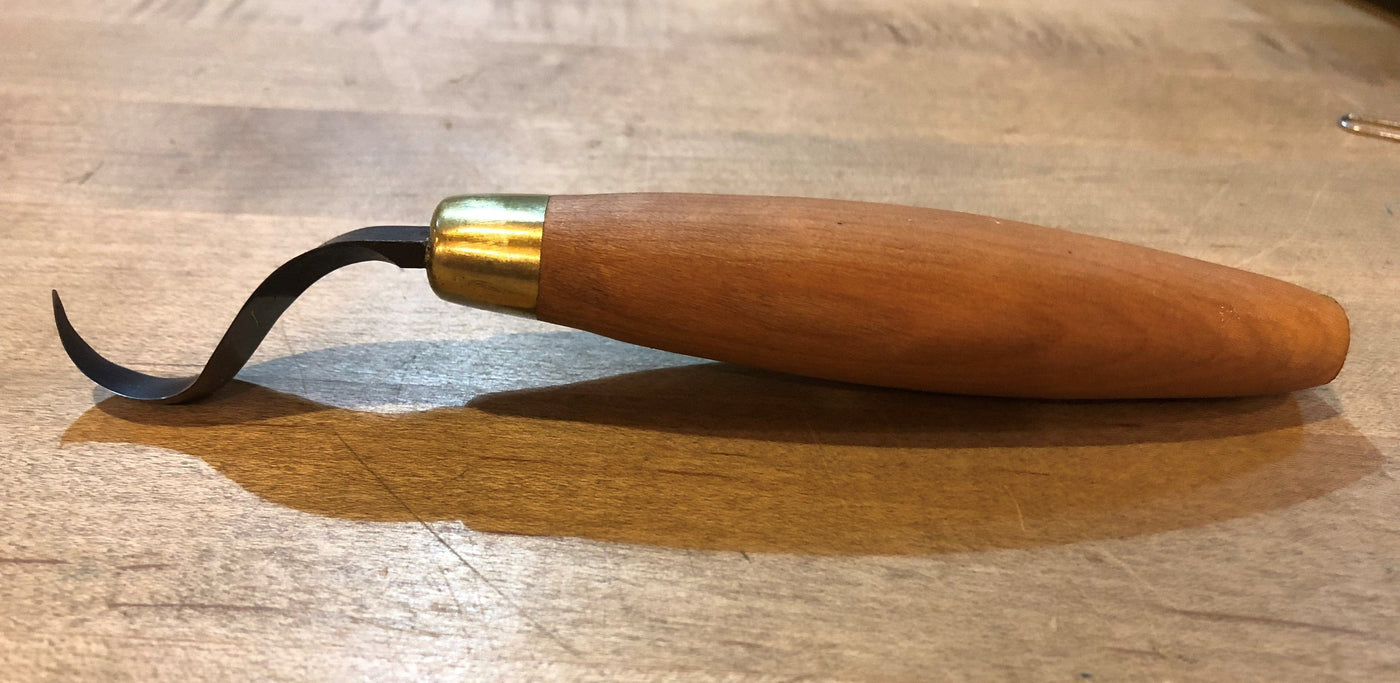 A FLE Sloyd Hook Knife, featuring a cherry hardwood handle and a curved high-carbon steel blade, rests on a wooden surface.
