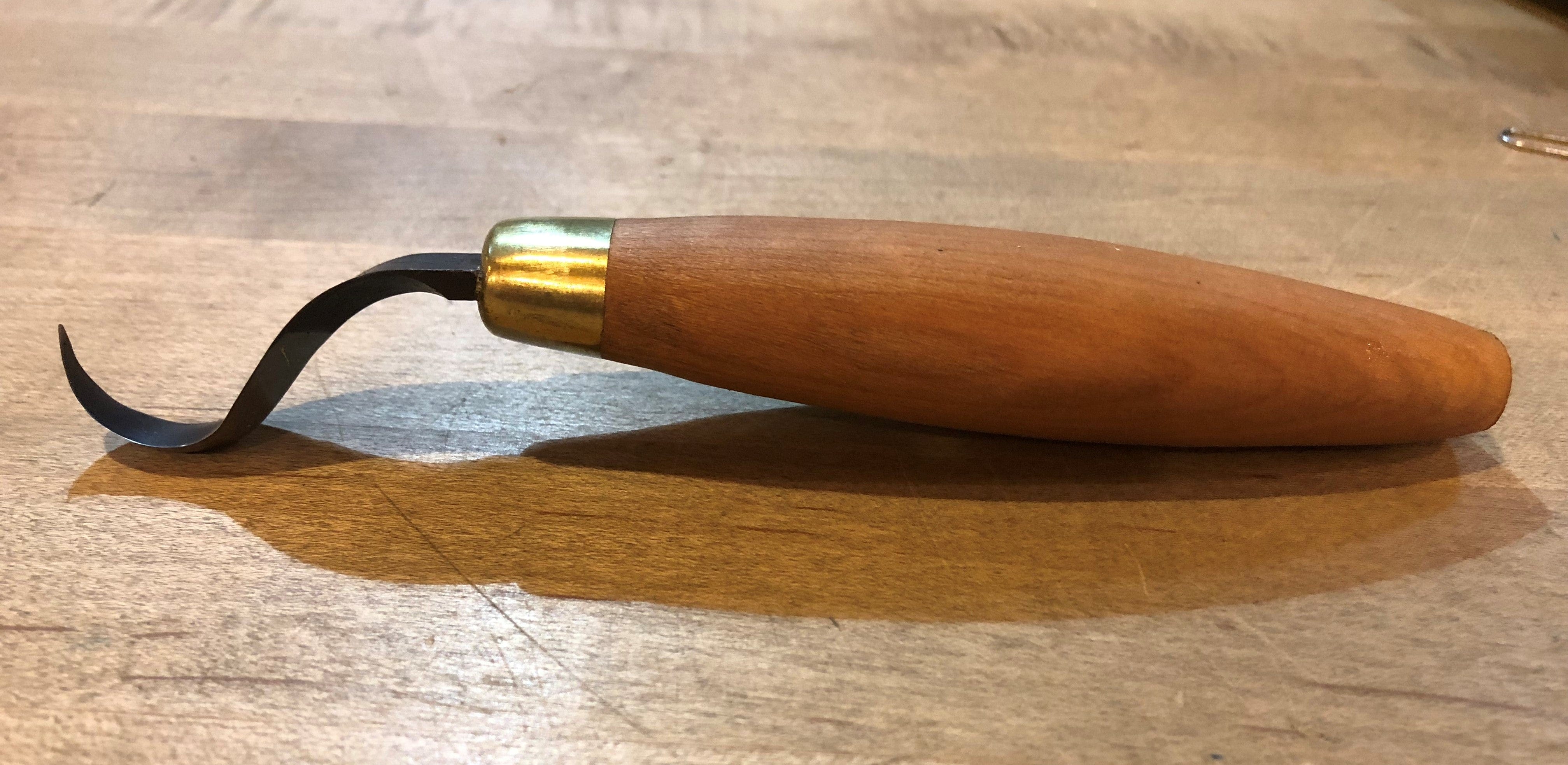 A FLE Sloyd Hook Knife, featuring a cherry hardwood handle and a curved high-carbon steel blade, rests on a wooden surface.
