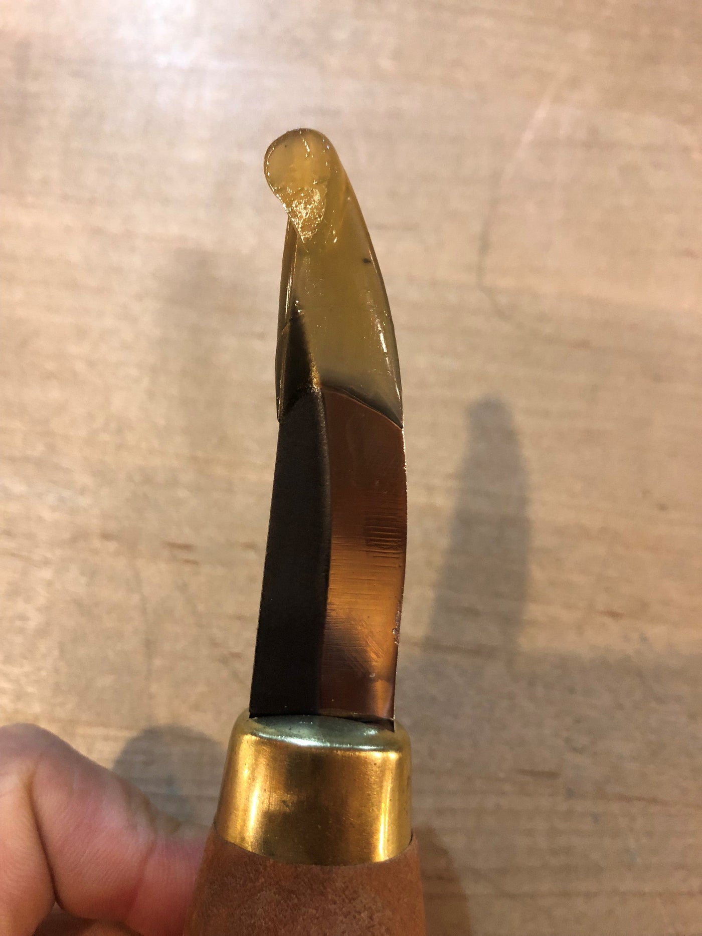 The FLE Stub Sloyd Knife, featuring a unique curved tip of translucent honey-like resin and a brass bolster, crafted from high-carbon steel, is displayed against a wood background.