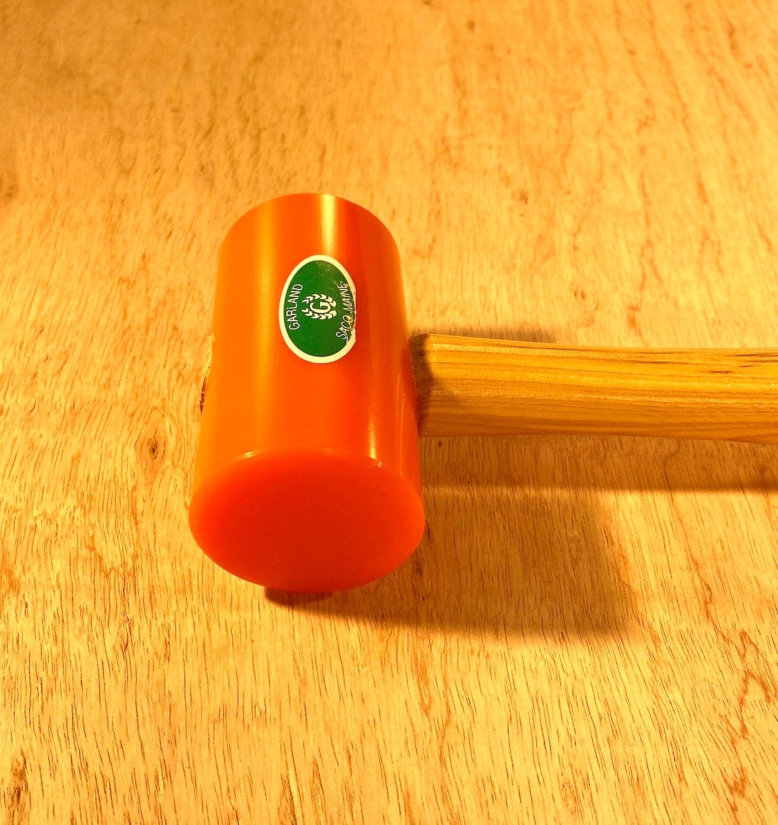 The Garland Elastomer Mallet by GAM, showcasing an orange body and a green logo on its head, rests elegantly on the wooden surface.