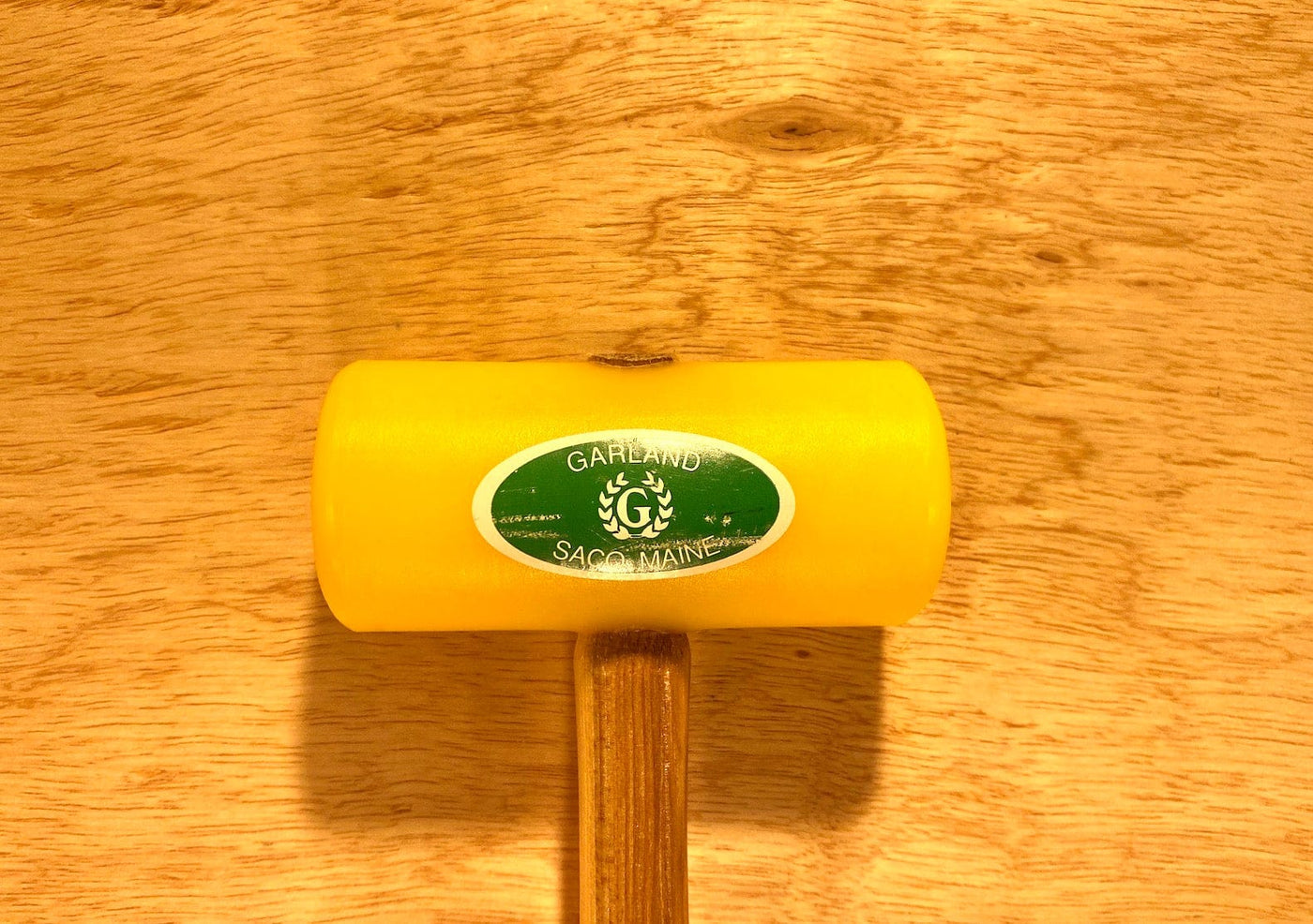 A GAM Garland Plastic Mallet, featuring a yellow head made from ultra-high-molecular-weight plastic for enhanced durability and easy cleaning, a wooden handle, and adorned with a green oval sticker that reads "Garland, Saco, Maine," is pictured on a wooden surface.