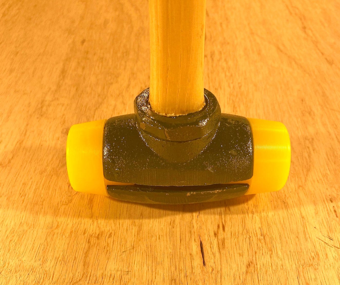 Close-up of a GAM Split Head Hammer Gar-Dur Face with a wooden handle resting on a wooden surface. The hammer's head, made of ultra-high-molecular-weight Gar-Dur plastic, is black with yellow ends.