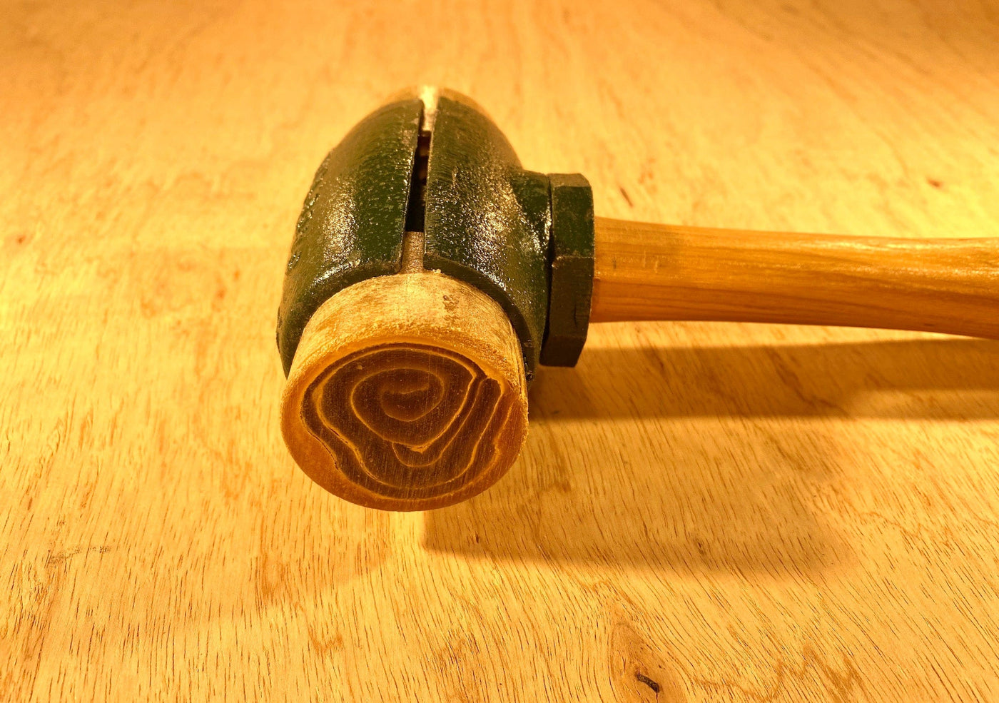 A close-up of a GAM Split Head Hammer Rawhide Face shows its wooden mallet head with visible end grain and concentric rings, plus a green painted section, lying on a light wood surface.