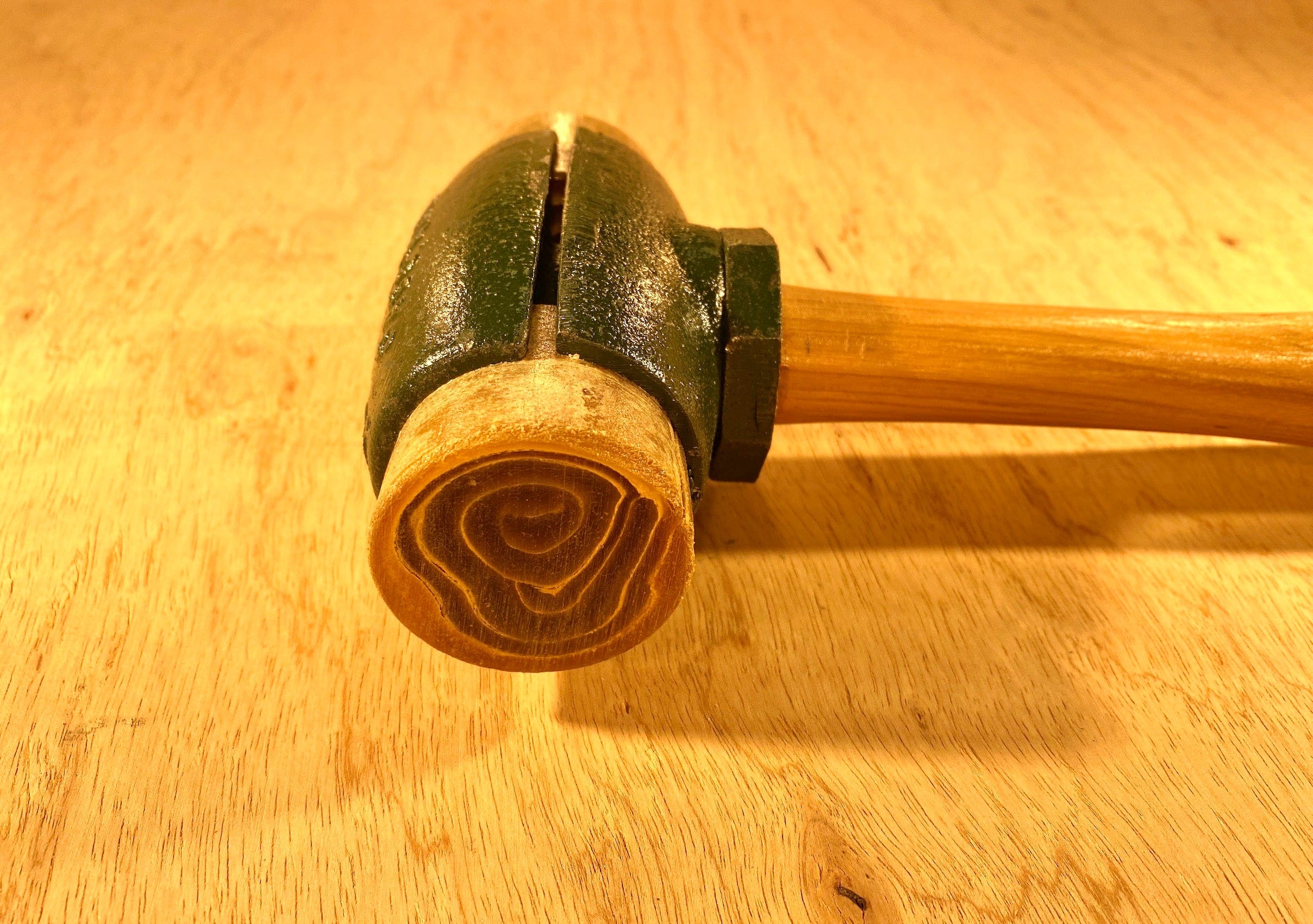 A close-up of a GAM Split Head Hammer Rawhide Face shows its wooden mallet head with visible end grain and concentric rings, plus a green painted section, lying on a light wood surface.