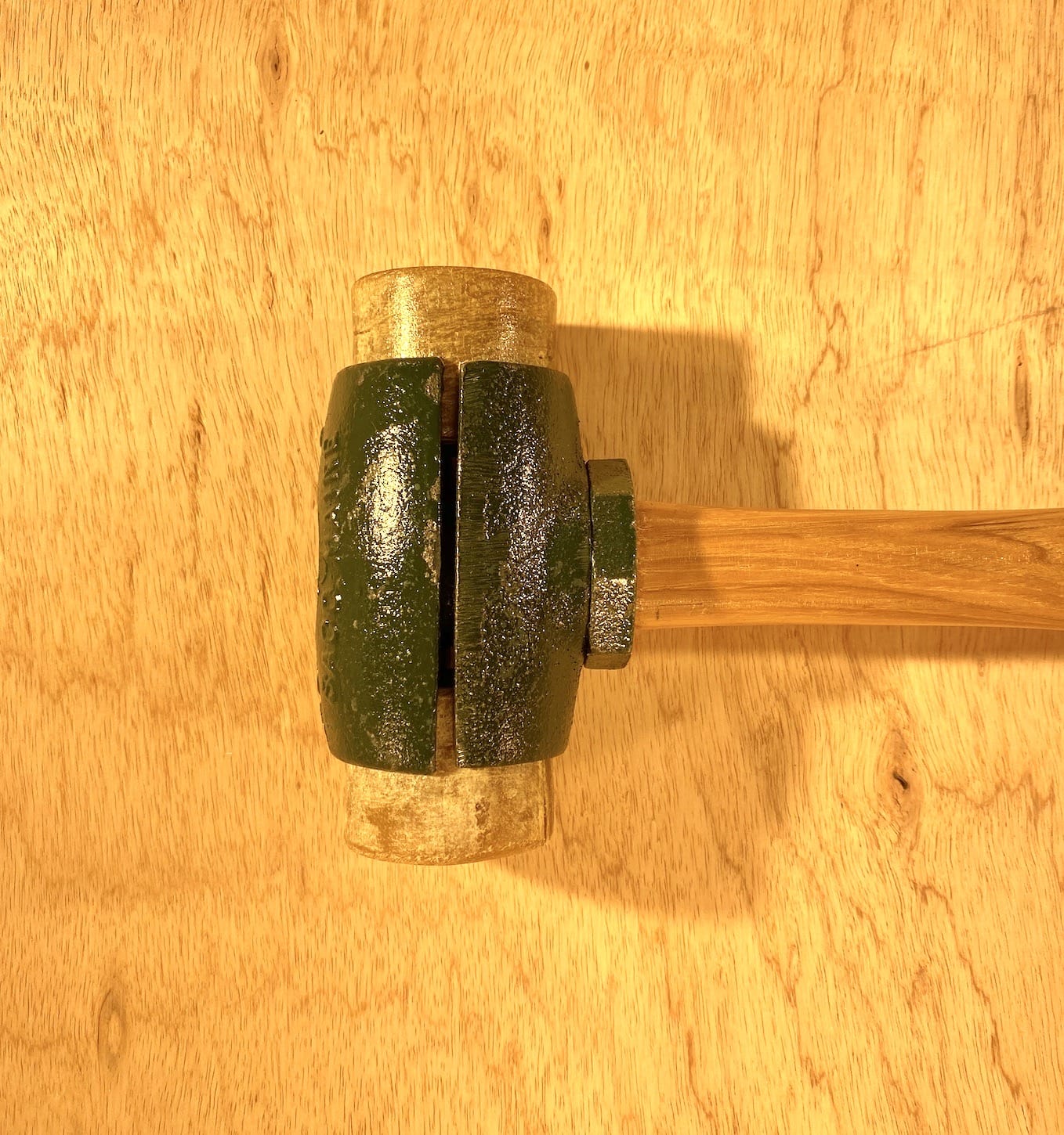 Close-up of a Split Head Hammer Rawhide Face from GAM, featuring a wooden handle and a green-painted metal head resting on a wooden surface.