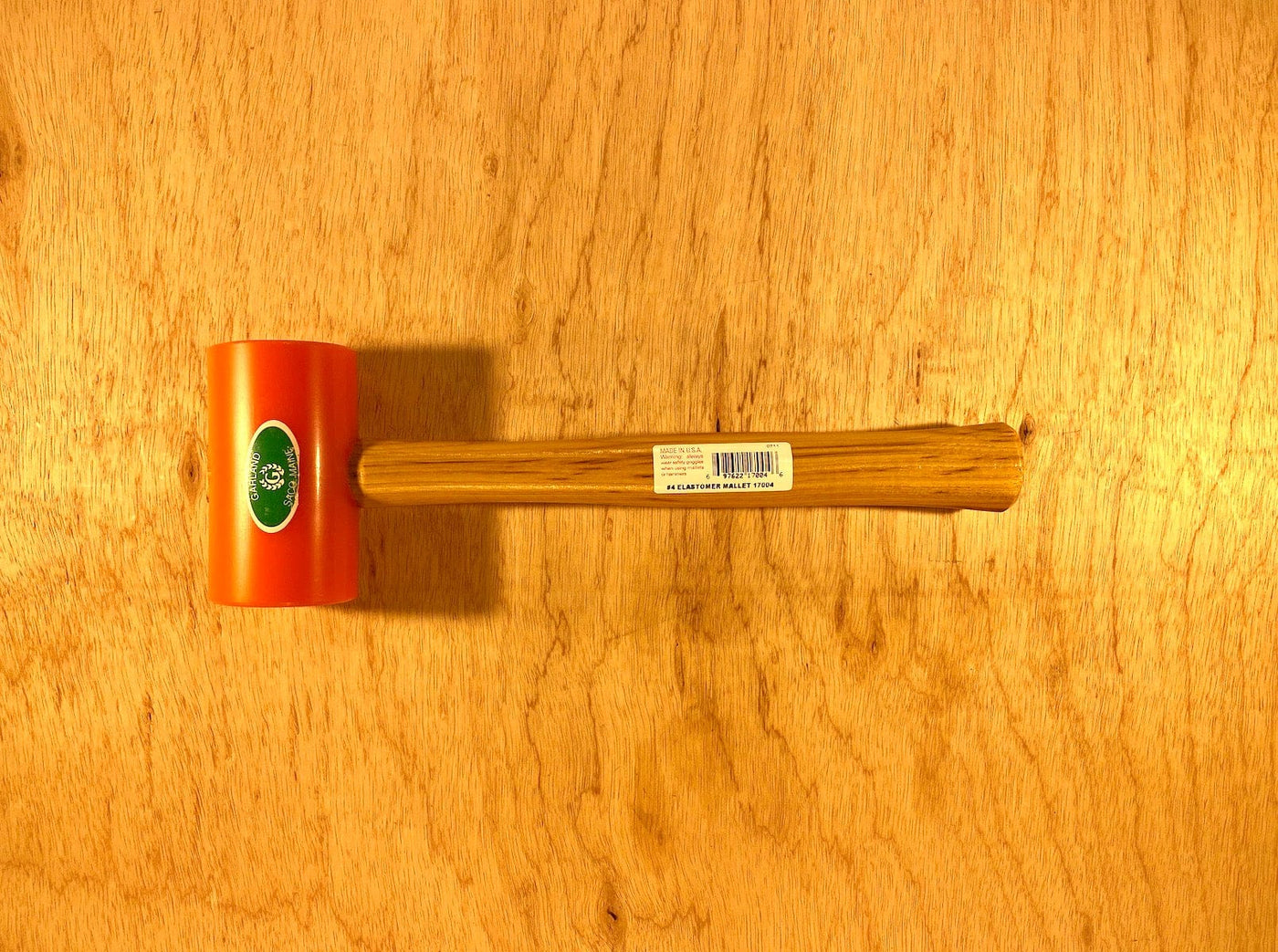 A Garland Elastomer Mallet with an orange head and wooden handle by GAM rests on a wooden surface, ideal for die-making applications.