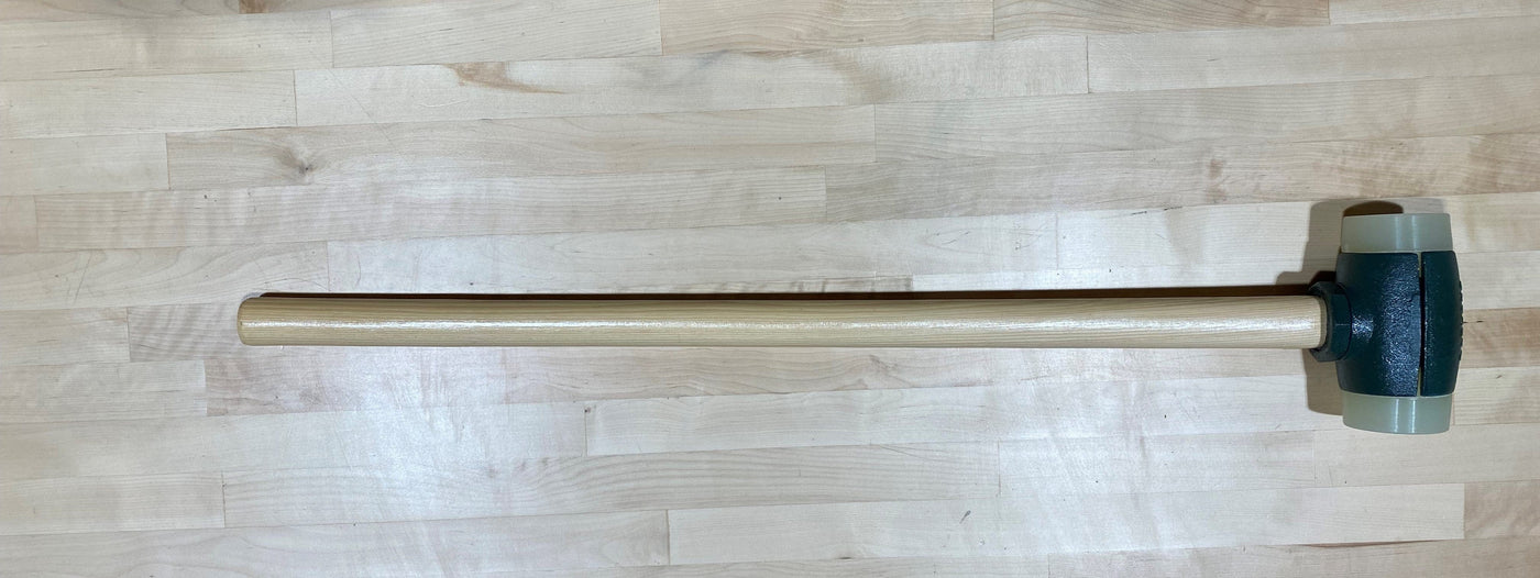 The Split Head Hammer Urethane Face by GAM, featuring a light wooden handle and dark rubber head, rests on a wooden surface.