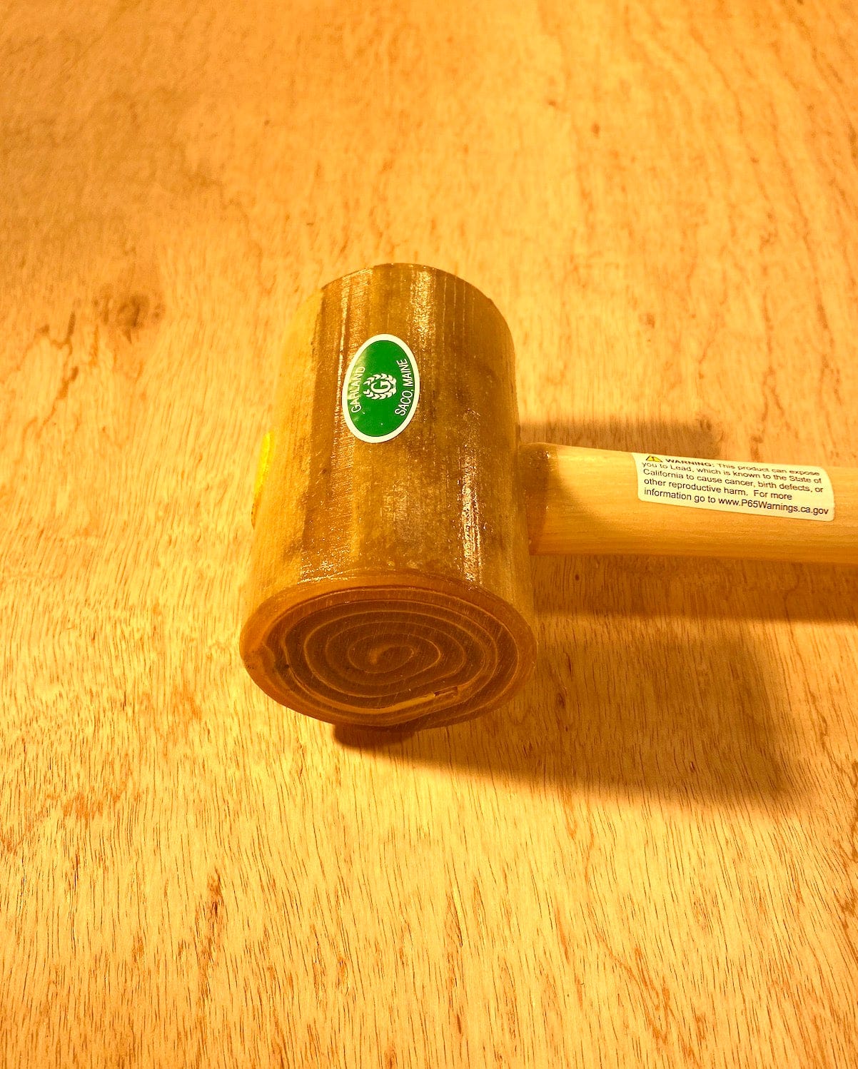 A wooden gavel with a circular design on the head and a green sticker is placed on a light wood surface, showcasing craftsmanship reminiscent of the Garland Weighted Rawhide Mallet by GAM.