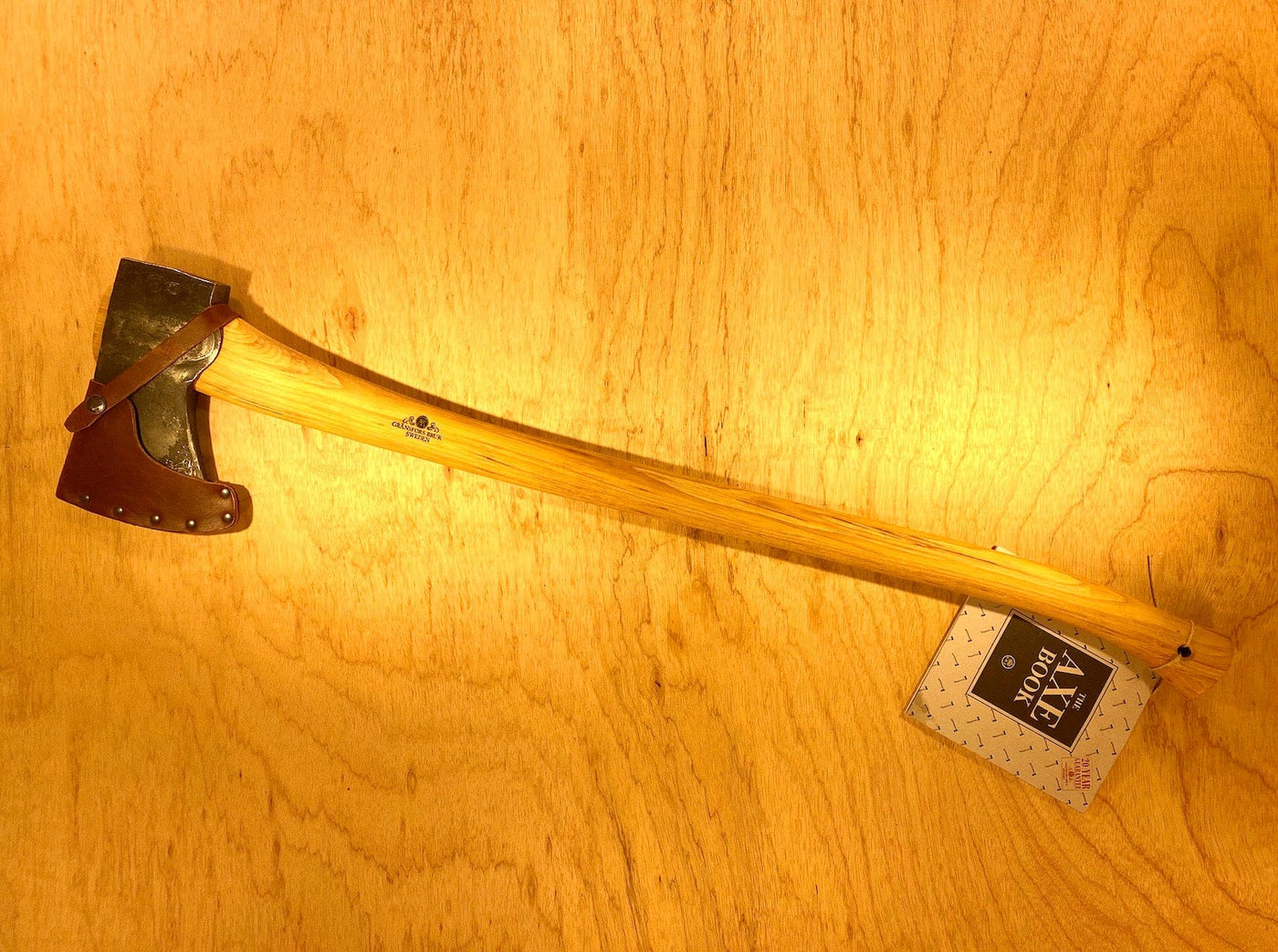The GRF Gransfors Bruk American Felling Axe with a 35-inch wooden handle and leather blade cover rests on a wooden surface, accompanied by a tag.