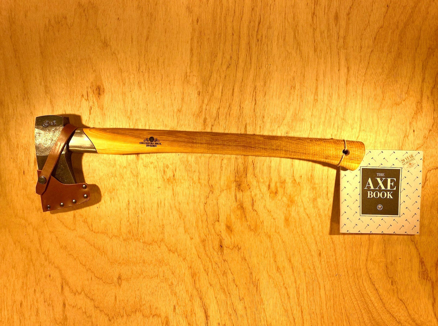 The Gransfors Bruk Small Splitting Axe 441 by GRF, featuring a hickory handle and a grain-leather sheath, is displayed on a wooden surface next to a booklet titled "The Axe Book.