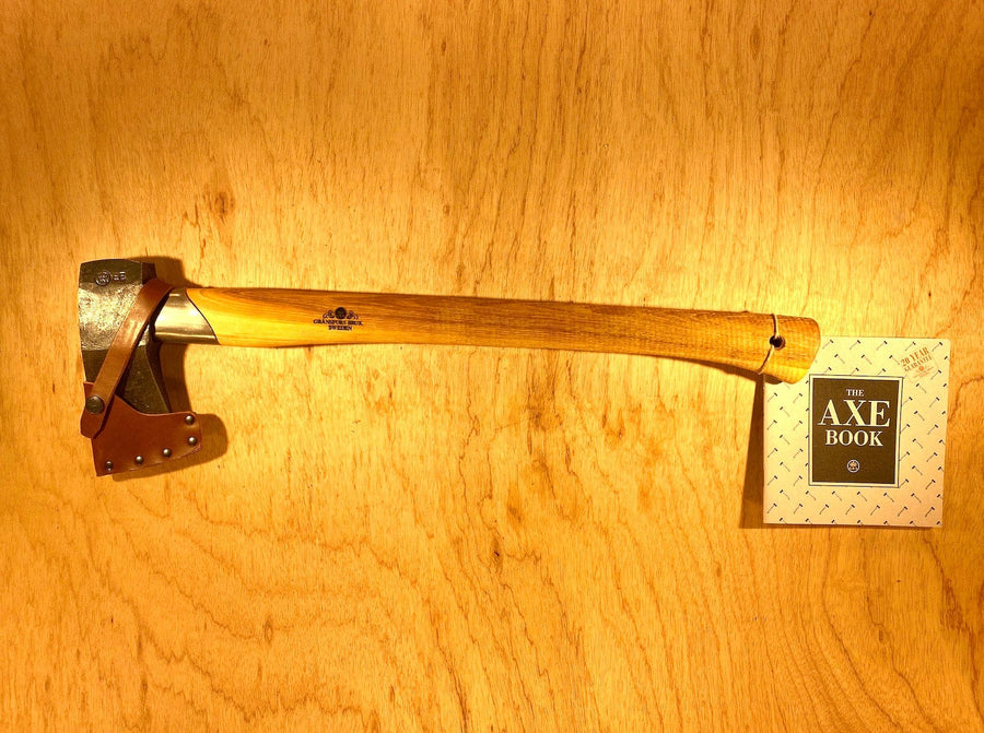 The Gransfors Bruk Small Splitting Axe 441 by GRF, featuring a hickory handle and a grain-leather sheath, is displayed on a wooden surface next to a booklet titled "The Axe Book.