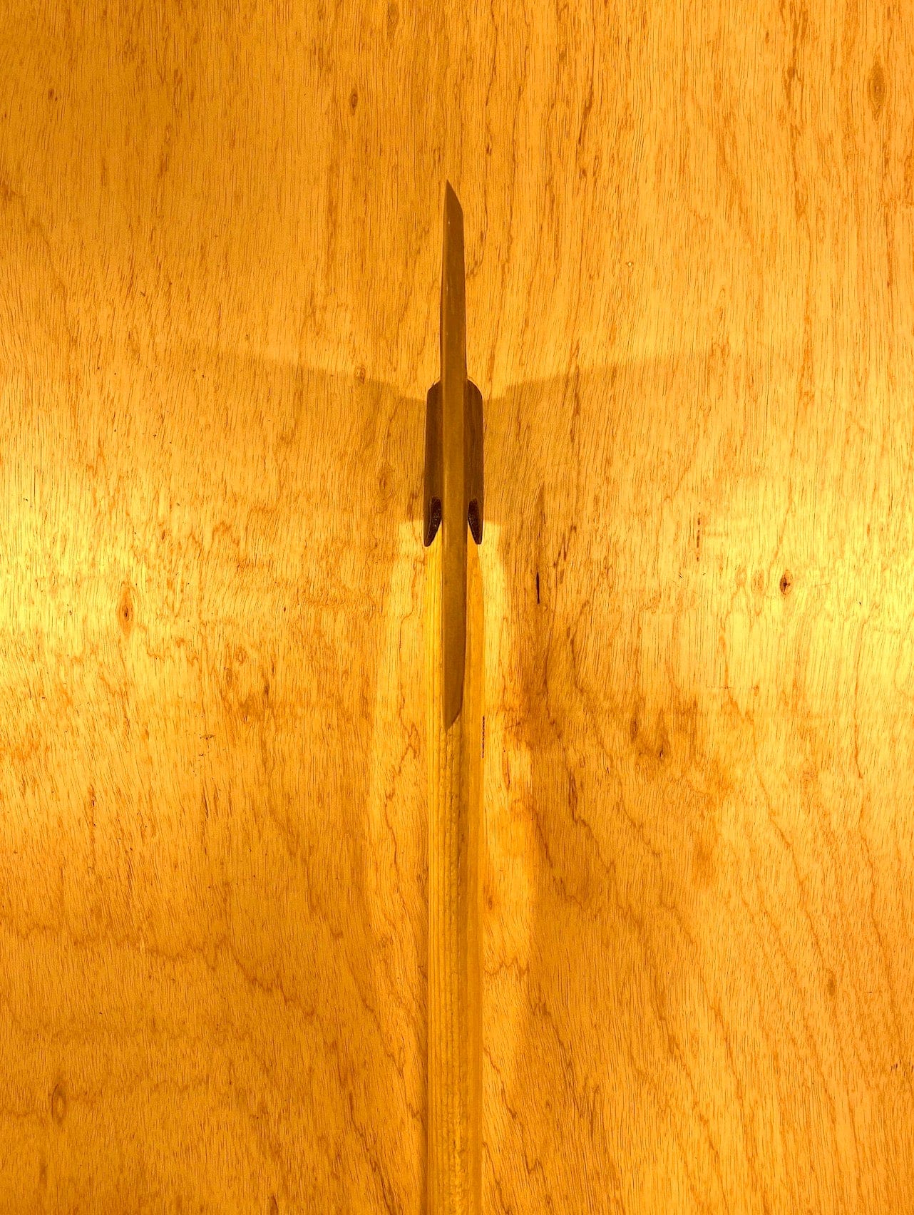 A Gransfors Bruk Broad Axe by GRF, expertly shaped for timber hewing, pierces a textured plywood surface, casting a shadow that resembles the silhouette of a rocket.