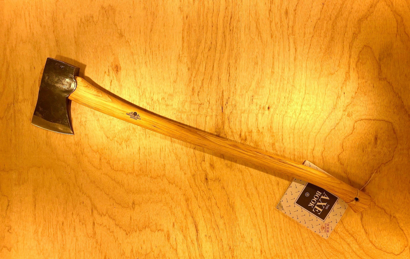 A Gransfors Bruk American Felling Axe with a wooden 35-inch handle and a steel head lies on a wooden surface, displaying the revered Swedish craftsmanship of GRF, accompanied by a tag labeled "The Best Made Co.