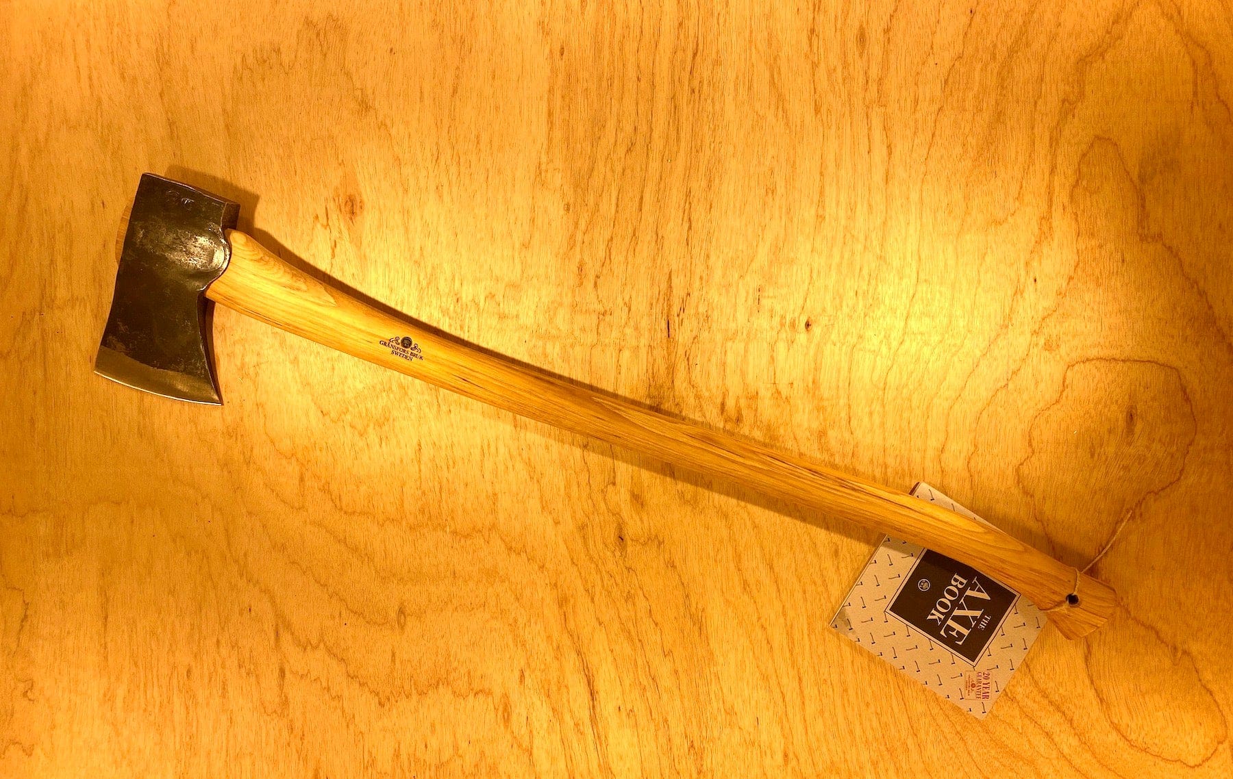 A Gransfors Bruk American Felling Axe with a wooden 35-inch handle and a steel head lies on a wooden surface, displaying the revered Swedish craftsmanship of GRF, accompanied by a tag labeled "The Best Made Co.
