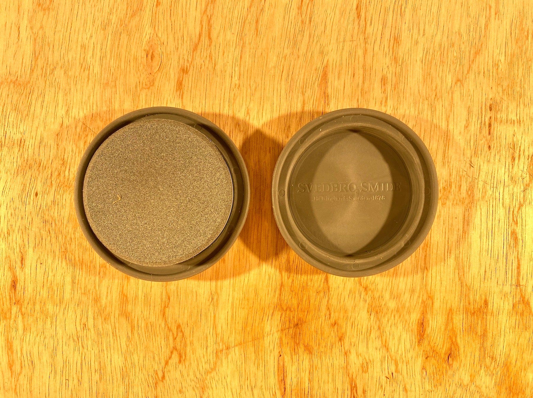 Two round, green GRF Ceramic Sharpening Stones with containers are on a wooden surface. The left container is open, revealing the stone; the right is closed with text on the lid, perfect for Gransfors Bruk fans aiming for precision sharpening.