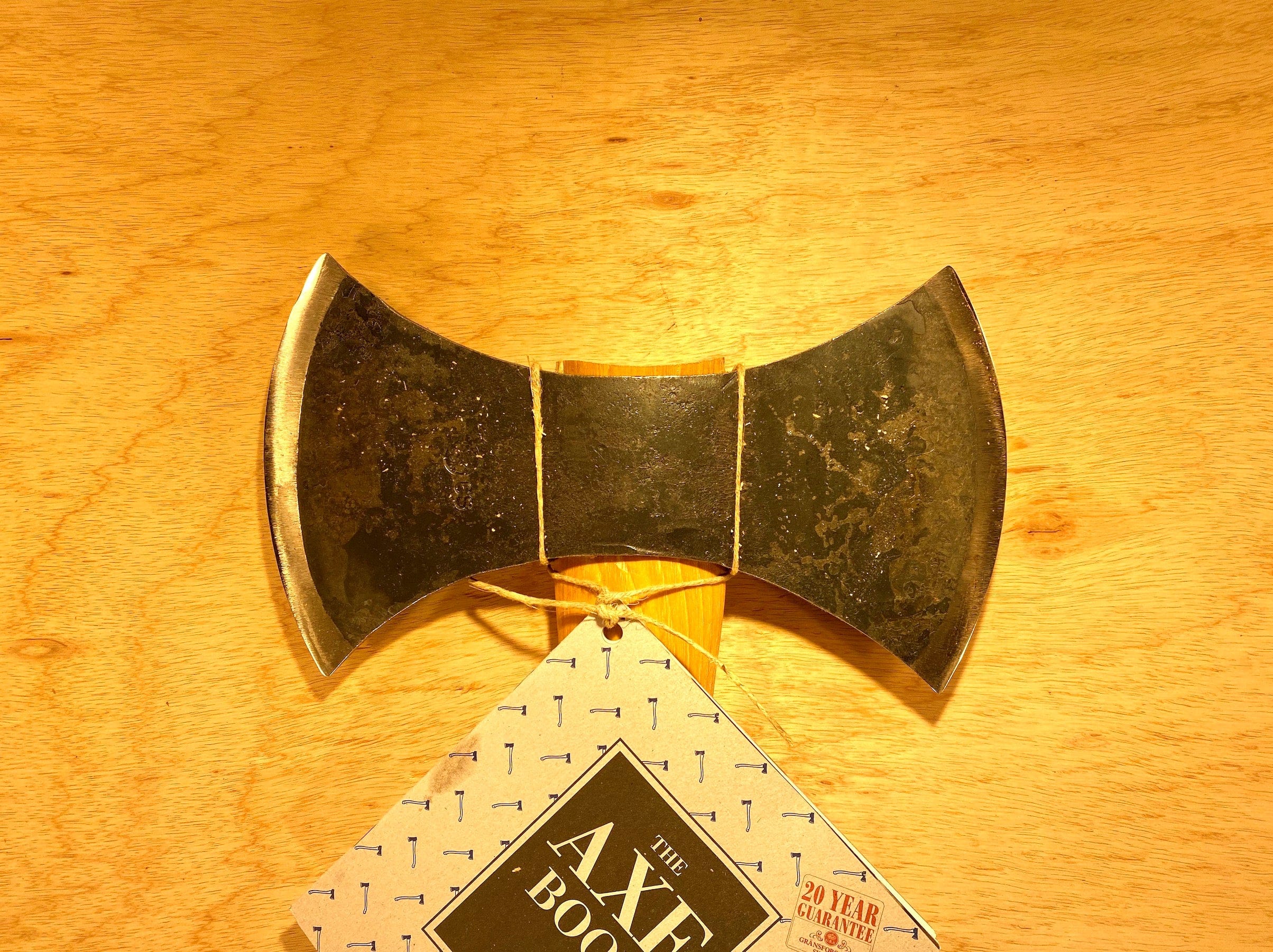 A Gransfors Bruk Double Bit Axe with a 35-inch handle from the brand GRF rests on a wooden surface beside "The Axe Book," accompanied by a 20-year guarantee sticker, making it perfect for enthusiasts of axe throwing.