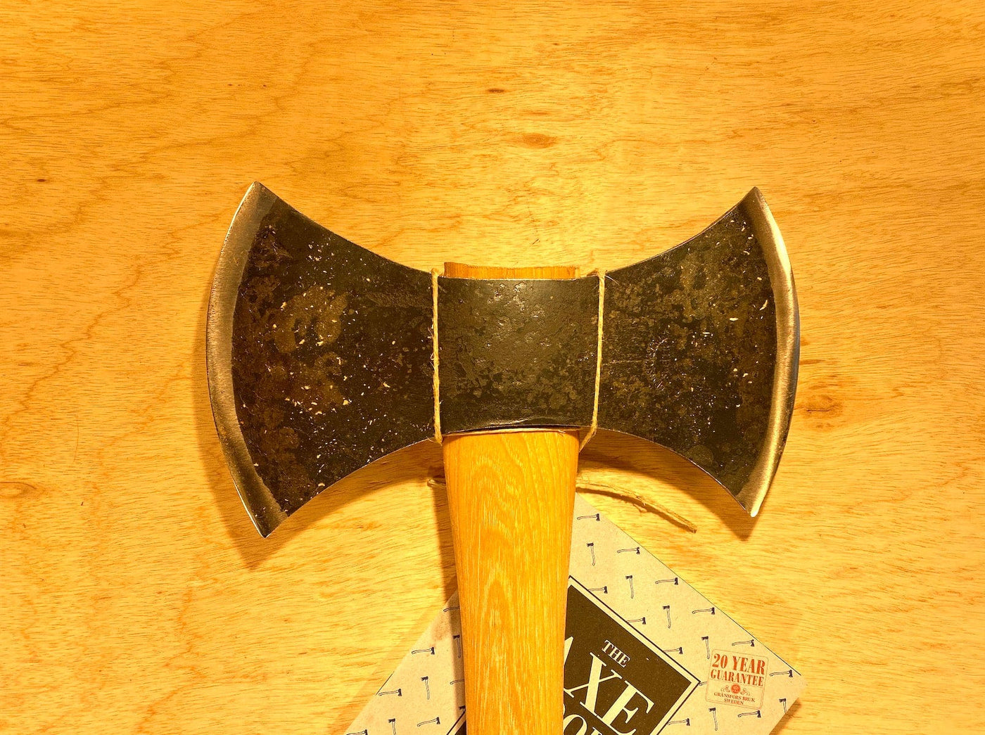 A close-up image of a GRF Gransfors Bruk Double Bit Axe with a 35-inch wooden handle resting on a wooden surface. A small booklet titled "The Axe Book" is partially visible underneath the axe, ideal for those interested in axe throwing techniques.