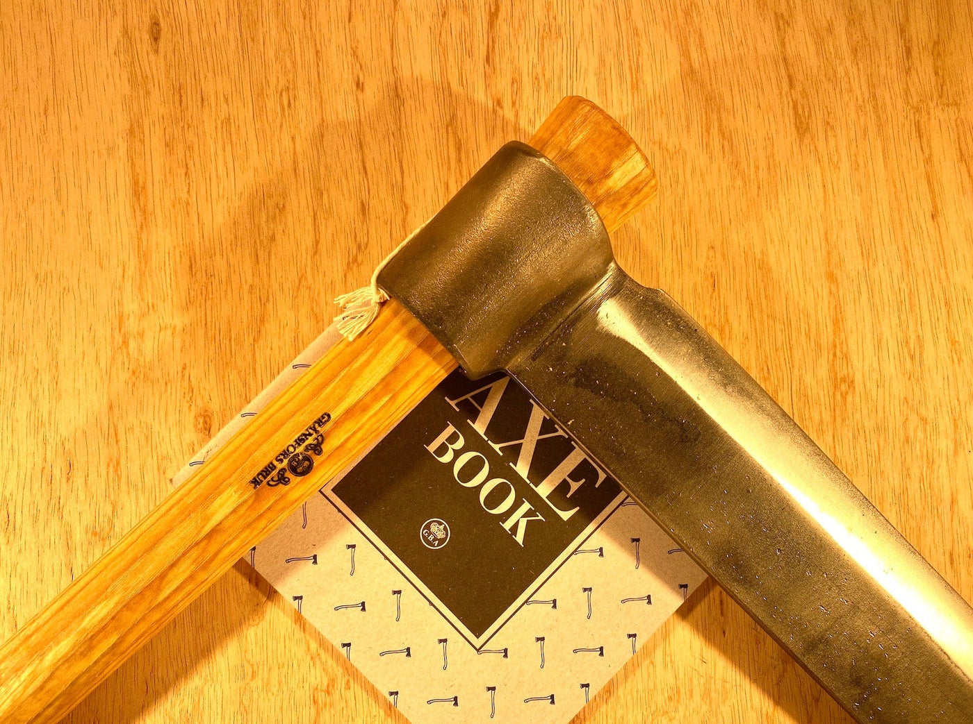 A Gränsfors Bruk Froe by GRF, featuring a wooden handle, rests atop a book titled "Axe Book" on a wooden surface, beautifully encapsulating the spirit of traditional woodworking.