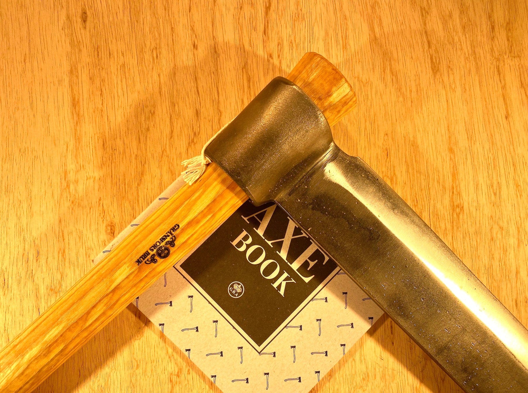 A Gränsfors Bruk Froe by GRF, featuring a wooden handle, rests atop a book titled "Axe Book" on a wooden surface, beautifully encapsulating the spirit of traditional woodworking.