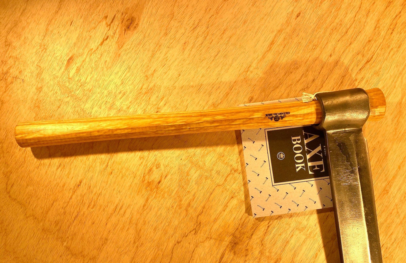 A GRF Gränsfors Bruk Froe and a wooden-handled axe rest on a wooden surface alongside a small booklet titled "Axe Book," making them ideal companions for your woodworking projects.