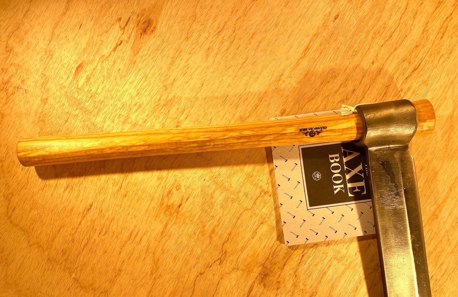 A GRF Gränsfors Bruk Froe and a wooden-handled axe rest on a wooden surface alongside a small booklet titled "Axe Book," making them ideal companions for your woodworking projects.