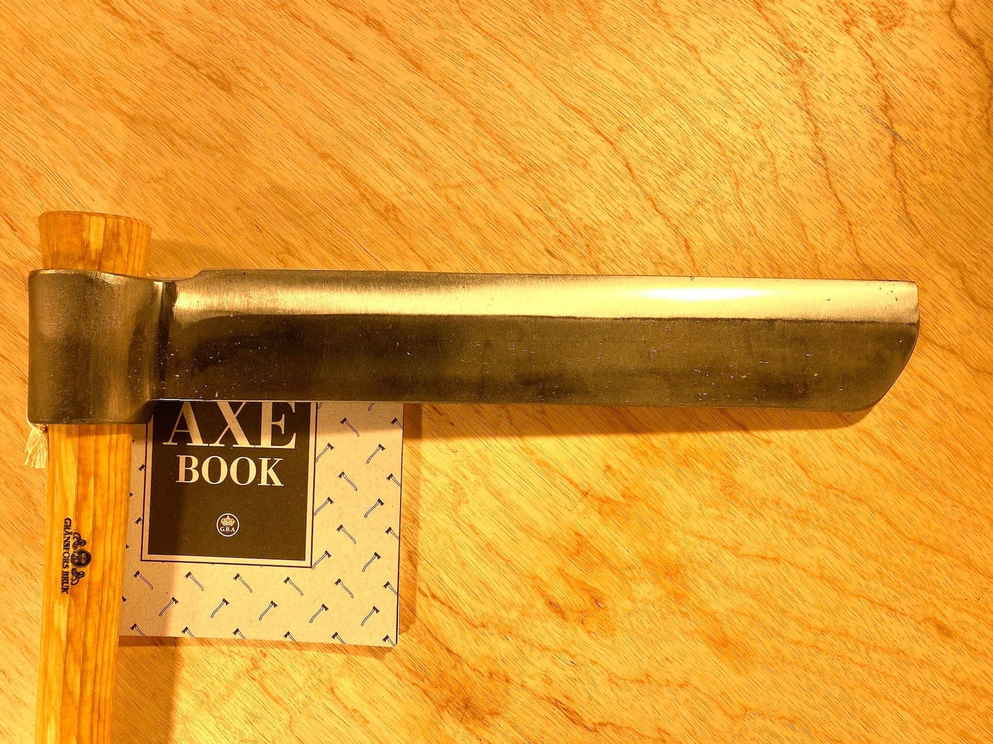 A Gränsfors Bruk Froe by GRF, featuring a wooden handle and ideal for woodworking, is placed on a small booklet titled "Axe Book" on a light wood surface.