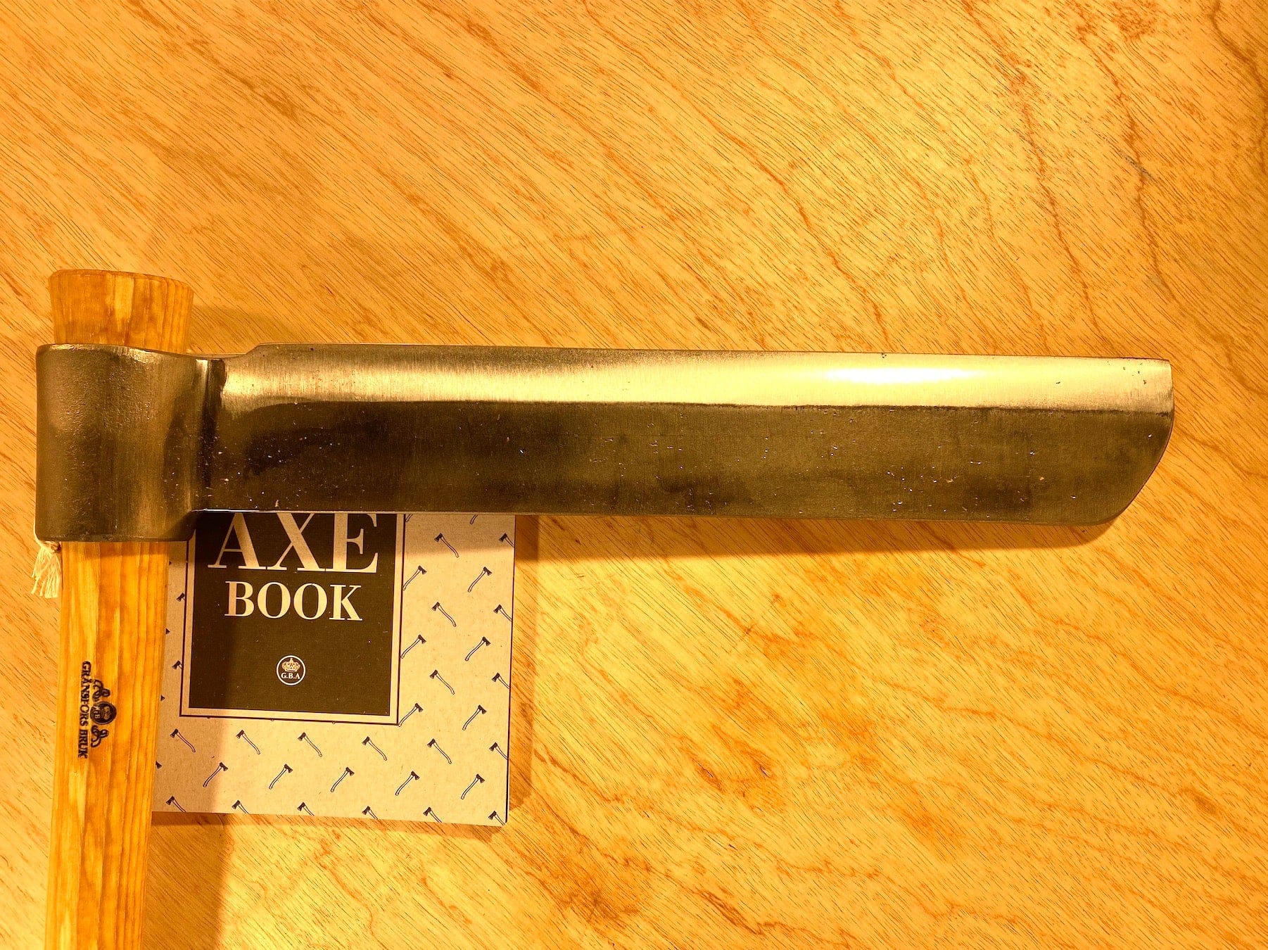 A Gränsfors Bruk Froe by GRF, featuring a wooden handle and ideal for woodworking, is placed on a small booklet titled "Axe Book" on a light wood surface.