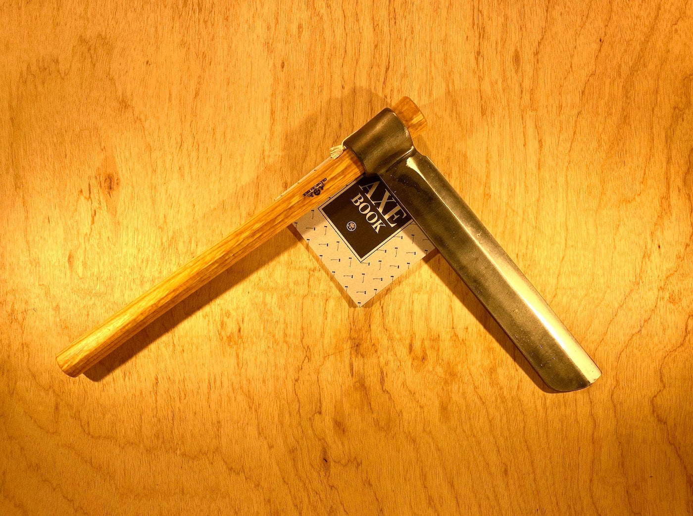 A Gränsfors Bruk Froe by GRF, featuring a wooden handle and metal blade, ideal for woodworking, rests on a wooden surface. A partially visible booklet titled "Axe Book" suggests techniques for wood-splitting and froe use.