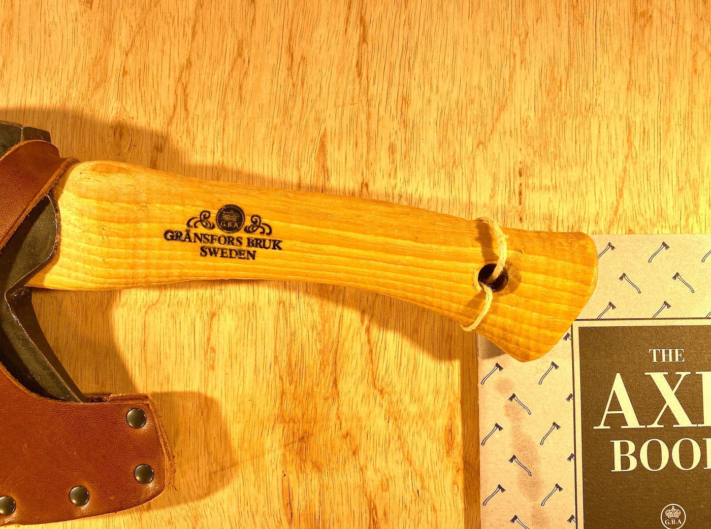A wooden-handled axe labeled "GRF, Sweden" rests on a wooden surface near a book titled "The Axe Book." This Gransfors Bruk Hand Hatchet 413 is perfect for firewood splitting or small tasks during scouting and camping adventures.