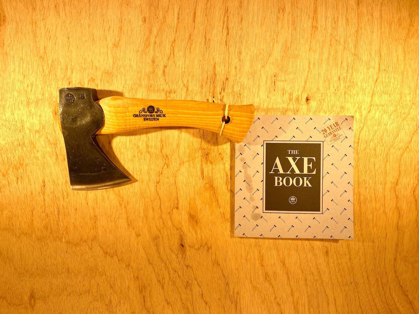 A Gransfors Bruk Hand Hatchet 413 by GRF with a wooden handle is displayed next to a book titled "The Axe Book" on a wooden surface, perfect for scouting and camping axe needs or firewood splitting.