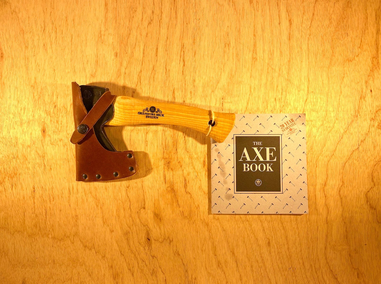 A GRF Gransfors Bruk Hand Hatchet 413 with a leather sheath is placed next to a book titled "The Axe Book" on a wooden surface, perfect for scouting and camping axe enthusiasts.