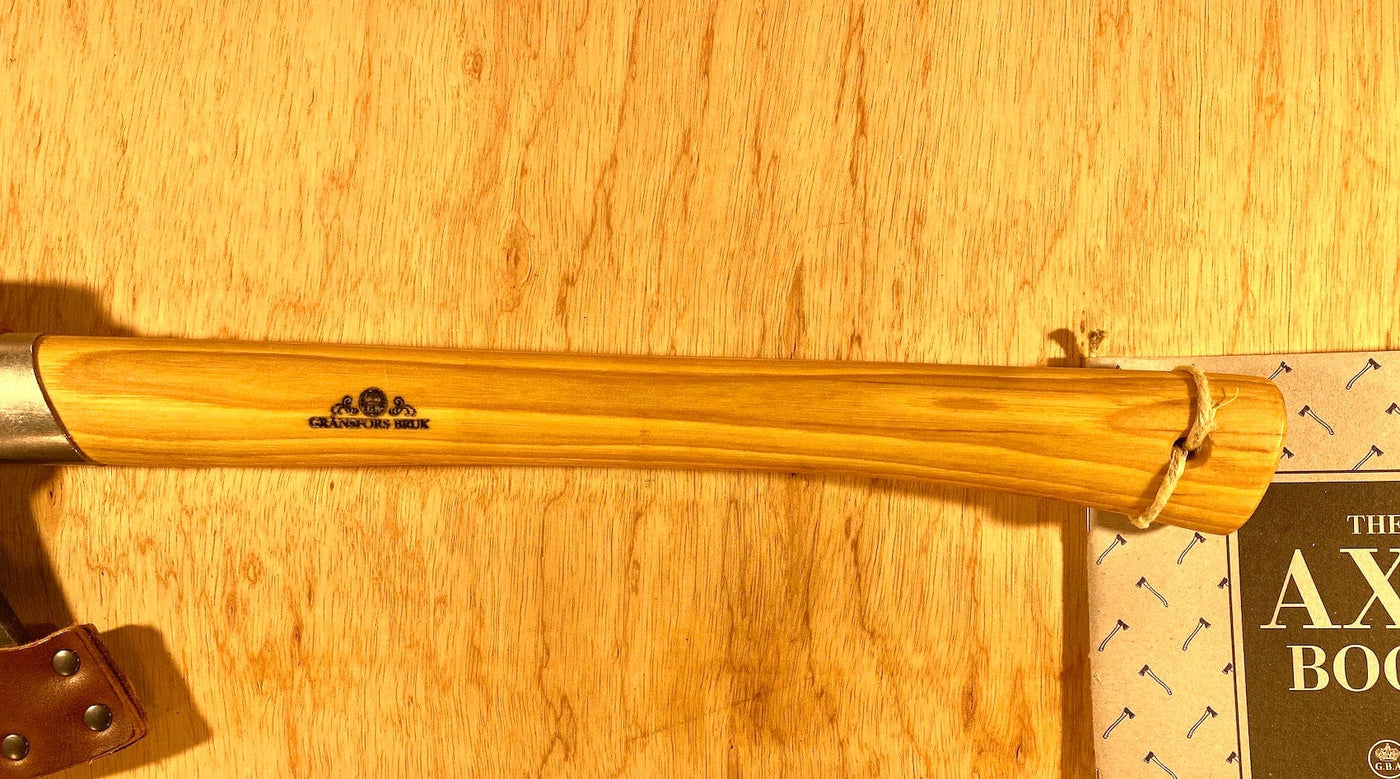A Gransfors Bruk Outdoor Axe by GRF with a wooden handle is beside "The Axe Book" on a wooden surface, inspired by Lars Fält and the teachings of the Swedish Armed Forces Survival School.