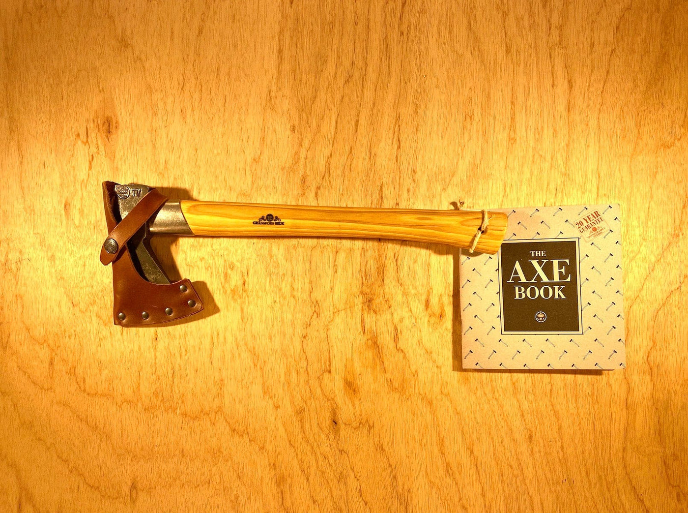 A GRF Gransfors Bruk Outdoor Axe with a wooden handle and leather sheath rests beside "The Axe Book" on a wooden surface, showcasing the expertise of survival expert Lars Fält from the Swedish Armed Forces Survival School.