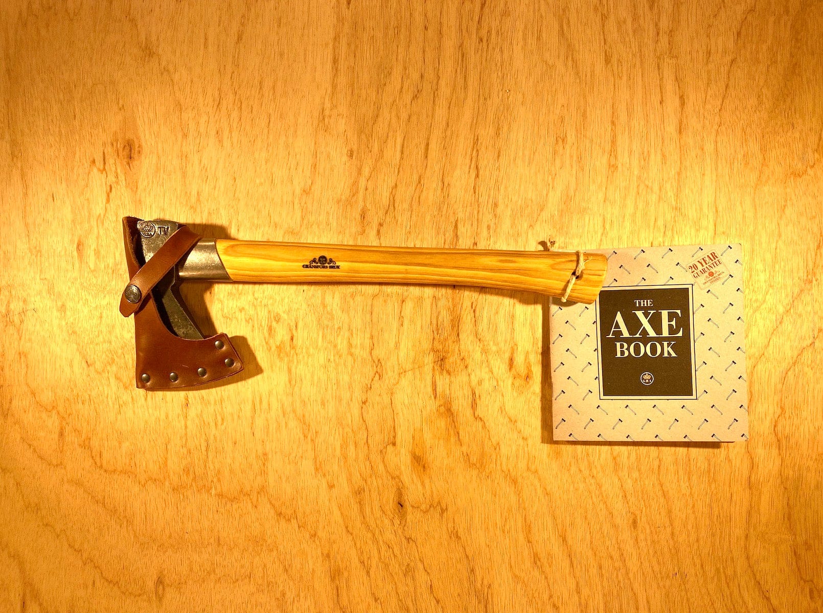A GRF Gransfors Bruk Outdoor Axe with a wooden handle and leather sheath rests beside "The Axe Book" on a wooden surface, showcasing the expertise of survival expert Lars Fält from the Swedish Armed Forces Survival School.