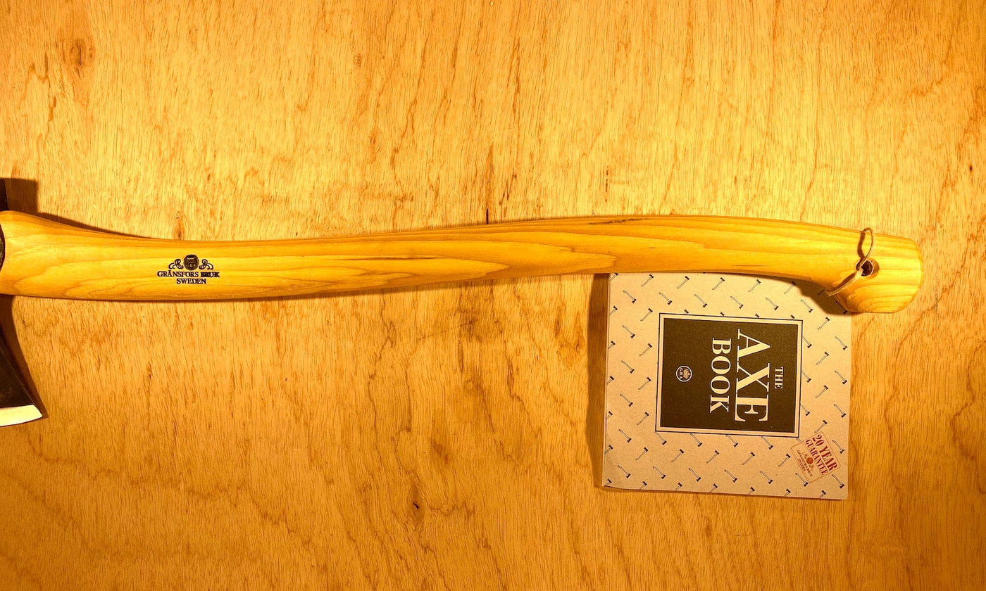 A GRF Gransfors Bruk Scandinavian Forest Axe, featuring a wooden handle and a black metal head, lies on a wooden surface next to a booklet titled "The Axe Book," showcasing it as an ideal professional forestry tool for felling and limbing.