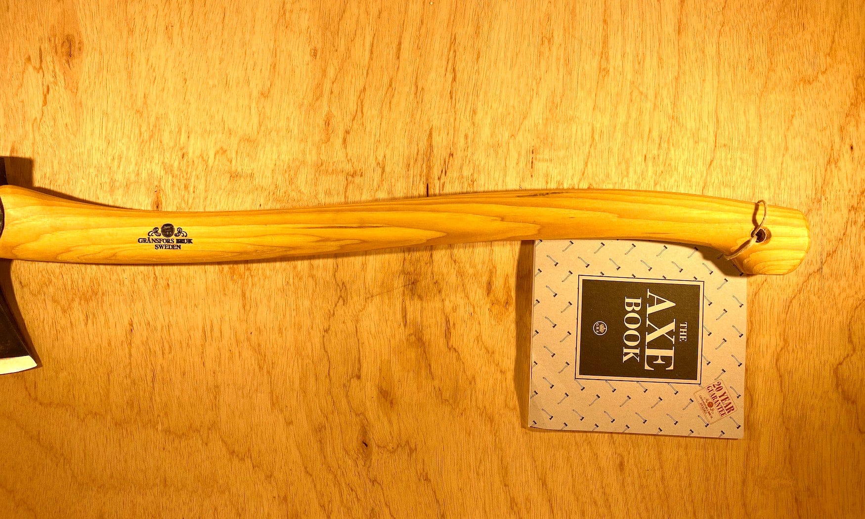 A GRF Gransfors Bruk Scandinavian Forest Axe, featuring a wooden handle and a black metal head, lies on a wooden surface next to a booklet titled "The Axe Book," showcasing it as an ideal professional forestry tool for felling and limbing.