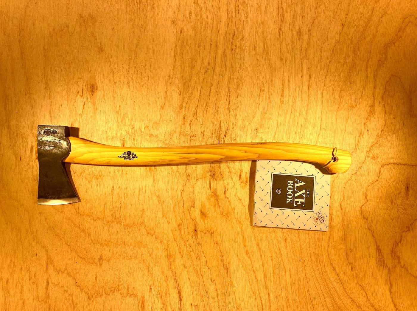 A Gransfors Bruk Scandinavian Forest Axe by GRF, featuring a wooden handle and metal head, is placed on a wooden surface. A tag labeled "AXE BOOK" is attached to the handle, identifying it as a professional forestry tool ideal for felling and limbing.