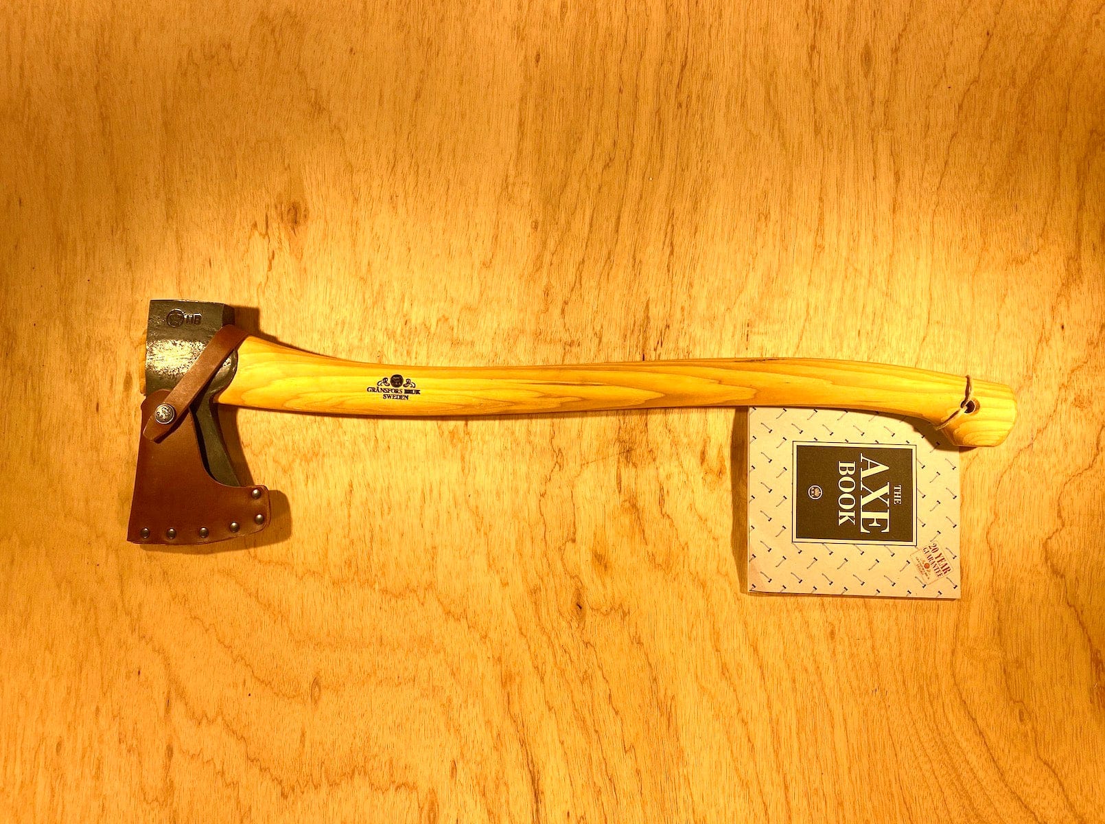 A Gransfors Bruk Scandinavian Forest Axe by GRF, featuring a wooden handle, rests on a wooden surface next to a book titled "Axe Book." The professional forestry tool's head is protected by a leather sheath, making it perfect for felling and limbing.
