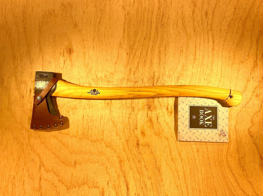 A Gransfors Bruk Scandinavian Forest Axe by GRF, featuring a wooden handle, rests on a wooden surface next to a book titled "Axe Book." The professional forestry tool's head is protected by a leather sheath, making it perfect for felling and limbing.