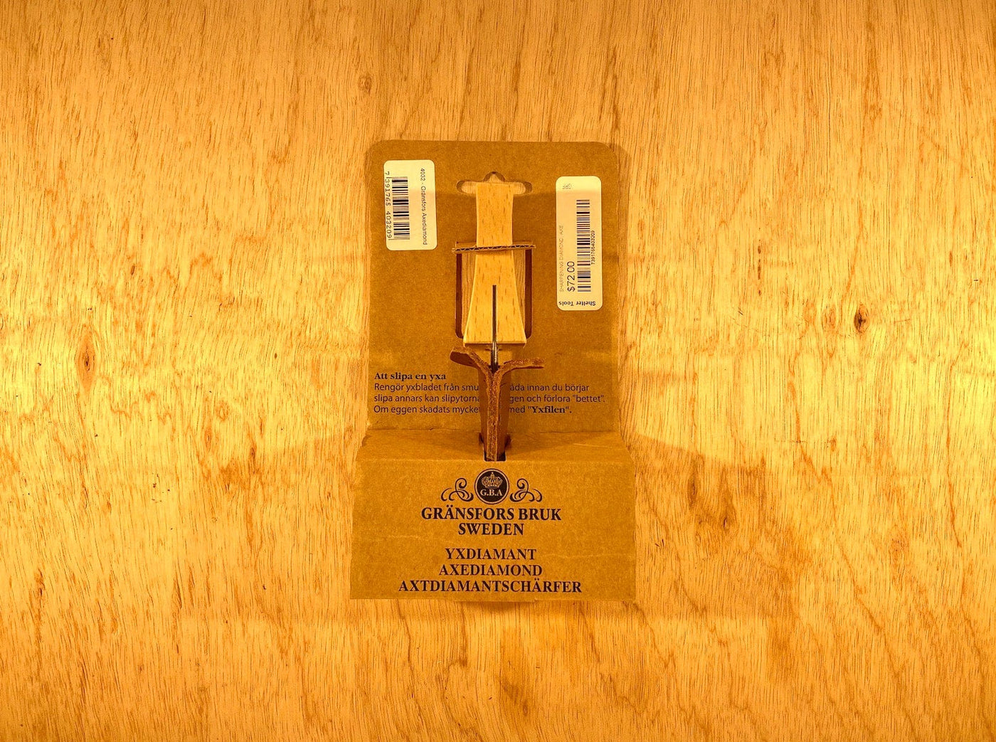 The Gransfors Bruk Sharpening Diamond File by GRF, featuring a wooden handle and both coarse and finer grit surfaces, is displayed on a cardboard backing with the brand "Gränsfors Bruk Sweden" and product details printed on it, all set against a rustic wooden background.