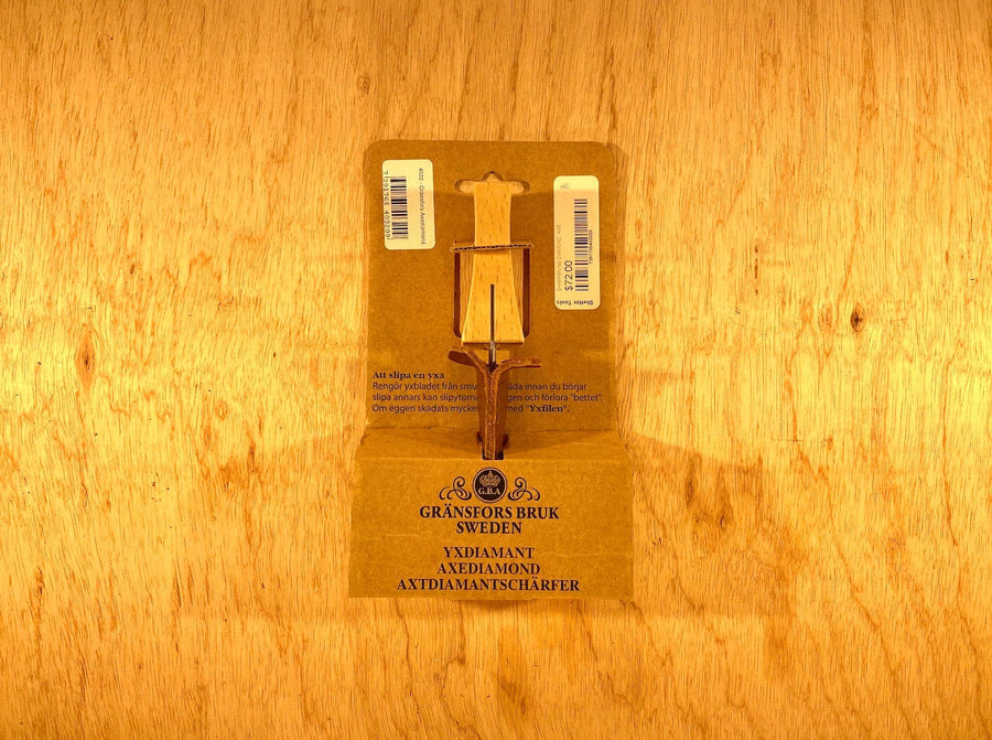 The Gransfors Bruk Sharpening Diamond File by GRF, featuring a wooden handle and both coarse and finer grit surfaces, is displayed on a cardboard backing with the brand "Gränsfors Bruk Sweden" and product details printed on it, all set against a rustic wooden background.