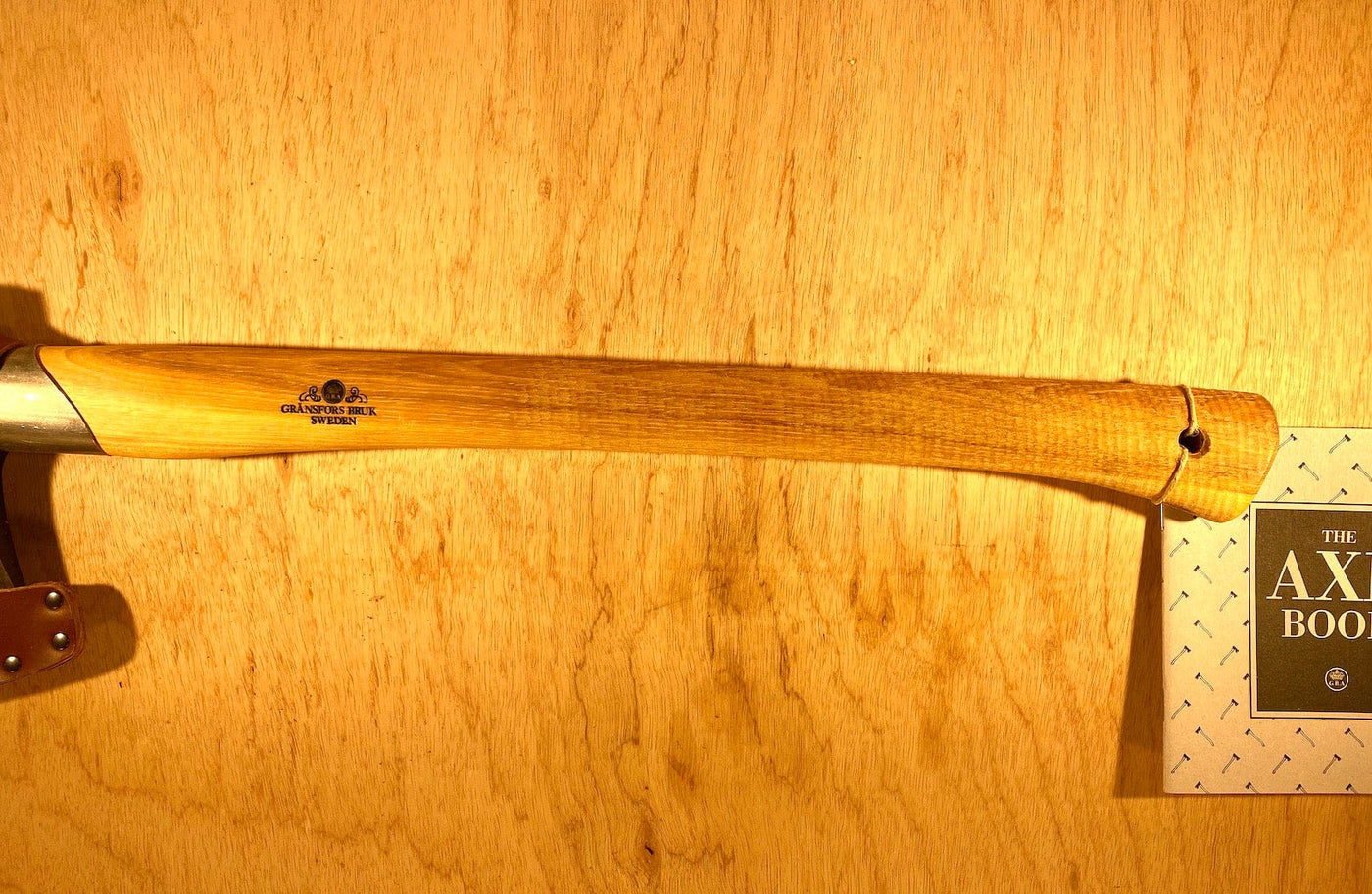 A Gransfors Bruk Small Splitting Axe 441 with a wooden handle, engraved with the brand name "GRF," featuring a grain-leather sheath, rests against a wooden backdrop next to a book titled "The Axe Book.