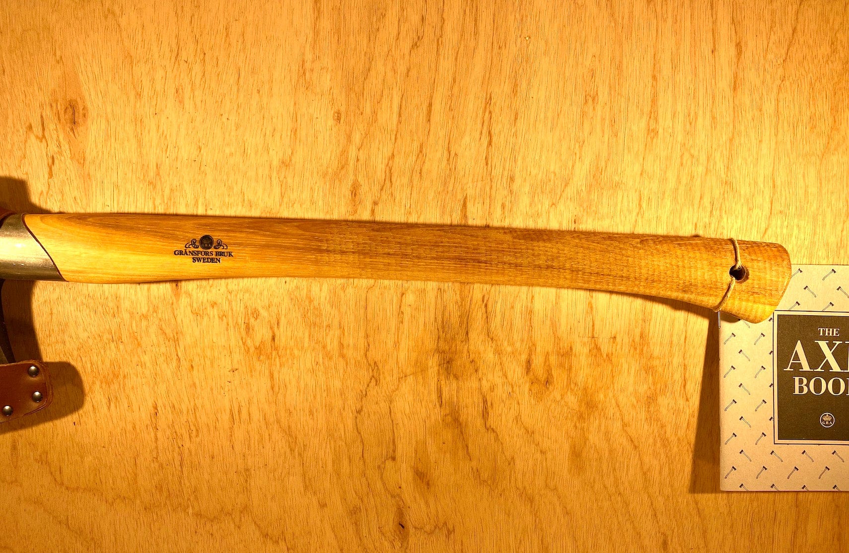 A Gransfors Bruk Small Splitting Axe 441 with a wooden handle, engraved with the brand name "GRF," featuring a grain-leather sheath, rests against a wooden backdrop next to a book titled "The Axe Book.
