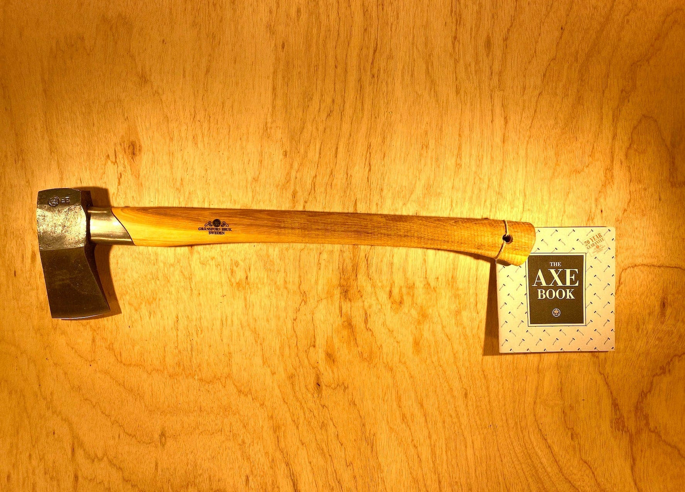 The Gransfors Bruk Small Splitting Axe 441 by GRF, featuring a sturdy hickory handle, is placed on a wooden surface. Attached to the handle is a booklet titled "The Axe Book.