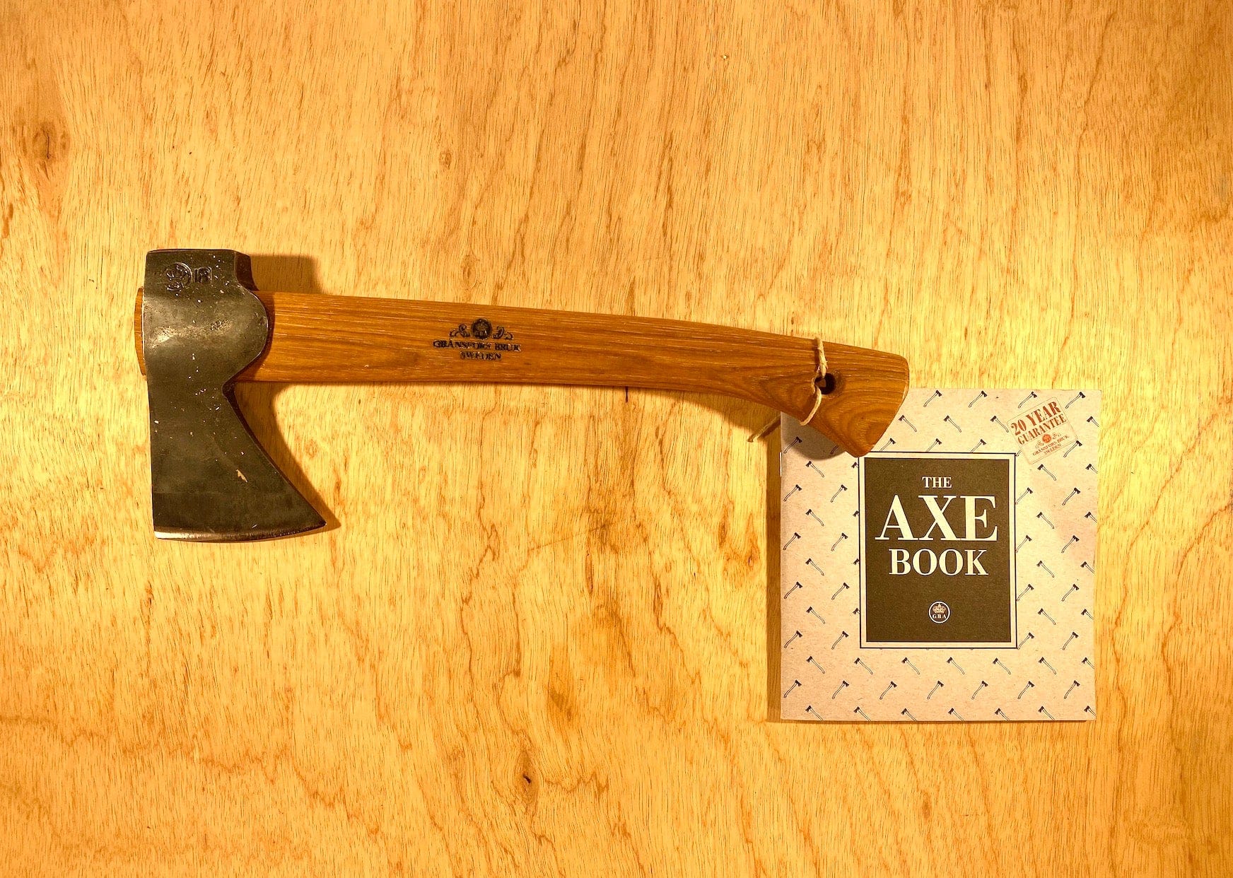 The Gransfors Bruk Wildlife Hatchet by GRF, featuring a wooden handle and "The Axe Book," rests on a wooden surface. Ideal for outdoor adventures, it blends traditional craftsmanship with timeless functionality.