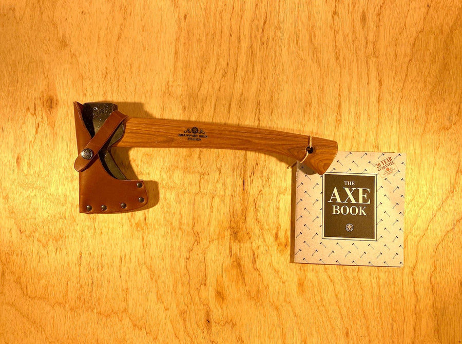A GRF Gransfors Bruk Wildlife Hatchet with a leather sheath lies on a wooden surface next to "The Axe Book." Its traditional craftsmanship is ideal for outdoor adventures.