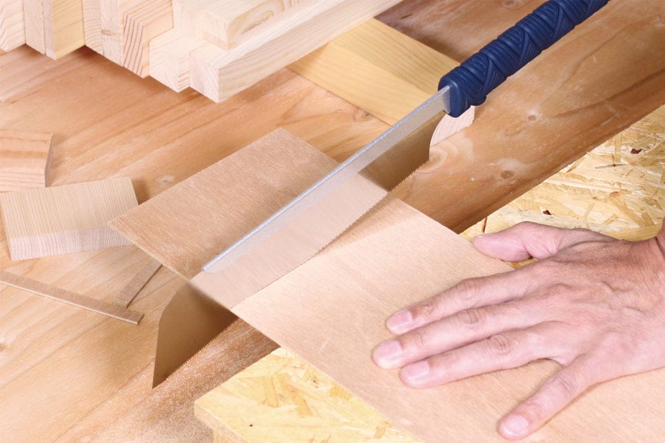 A person uses the BBT Dozuki Cross Cut Razorsaw to make precise cuts in a thin wooden board on a workbench, surrounded by wood pieces and planks, demonstrating expert carpentry.