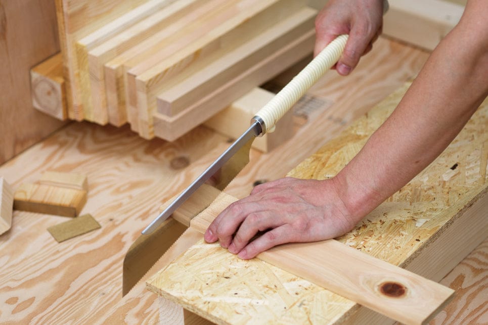 A person uses a BBT Dozuki Cross Cut Razorsaw with a white handle to cut wood on a workbench, with stacks of lumber and wood panels nearby for finishing or carpentry projects.