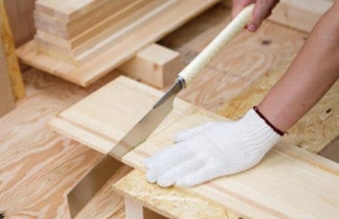 A person wearing a white glove uses the BBT Super Hard Cross Cut Razorsaw 270MM 13TPI to saw a wooden board, with stacked planks nearby.